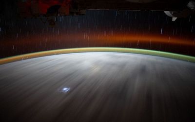 A long-exposure photo taken from the International Space Station shows airglow as bands of green and red curving around Earth. A flash of lightning appears near the bottom.