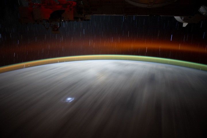 A long-exposure photo taken from the International Space Station shows airglow as bands of green and red curving around Earth. A flash of lightning appears near the bottom.